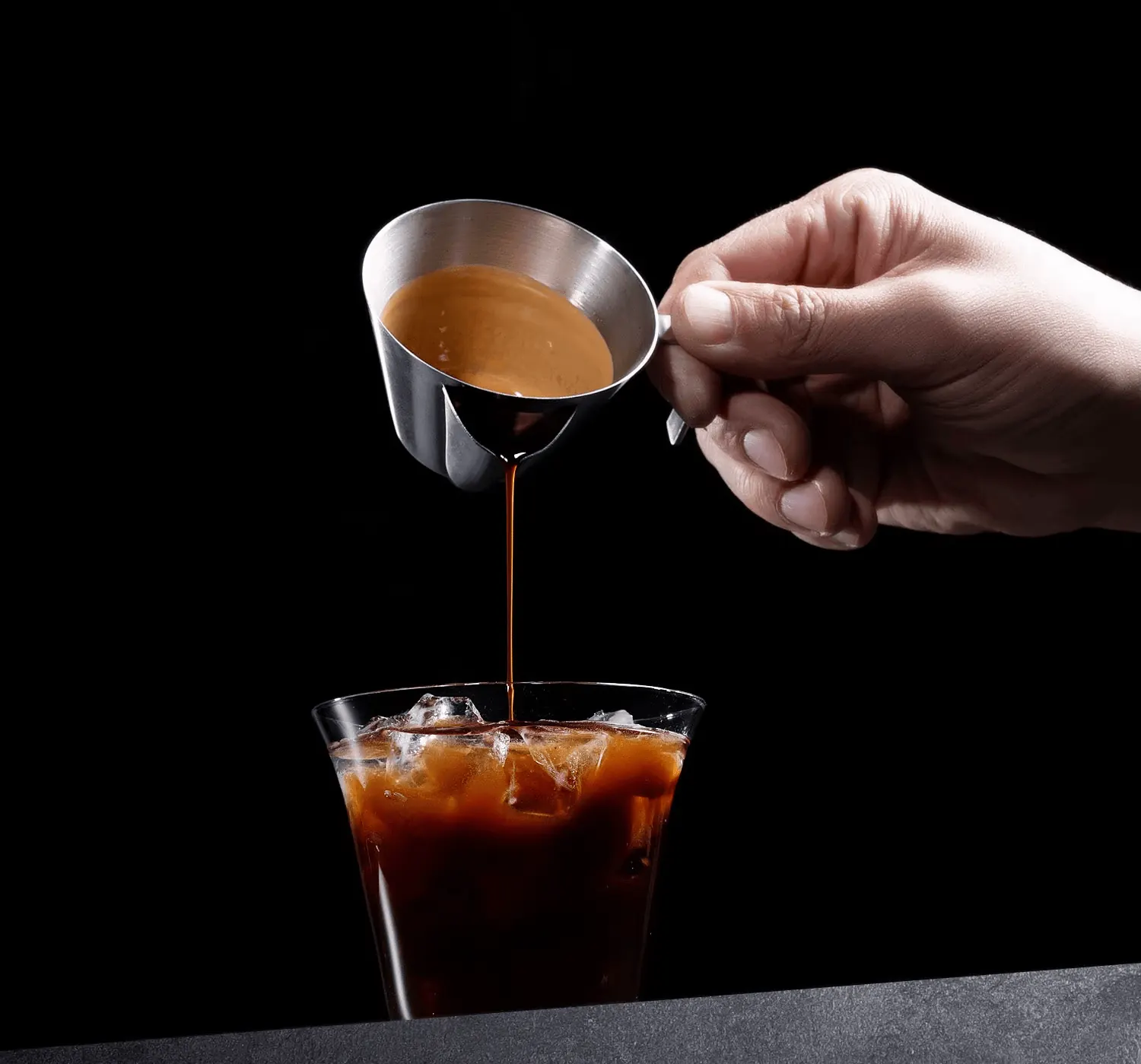 Čisté espresso stéká z nerezové konvičky do sklenice s ledem, crema zůstává v konvičce